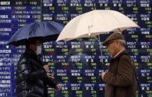 Pedestrians holding umbrellas walk past an electronic board showing stock prices outside a brokerage in Tokyo