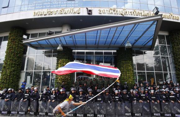 An anti-government protester waves a national flag in front of riot police officers and soldiers guarding the entrance of the National Broadcast Services of Thailand (NBT) television station in Bangkok May 9, 2014. CREDIT: REUTERS/ATHIT PERAWONGMETHA