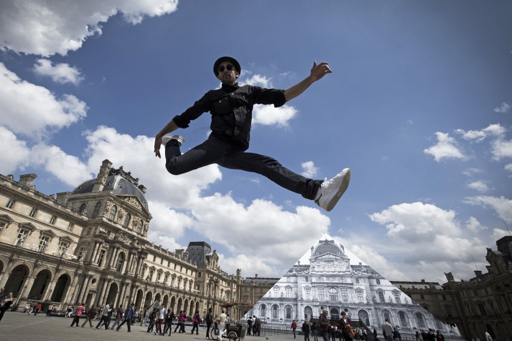 French artist and photographer JR jumps in front the Louvre pyramid after recovering it 'with a surprising anamorphic image', according to the museum in Paris on May 25, 2016. This recovering introduces the opening of JR exhibition 'Contemporary art JR at the Louvre' will run from May 25 to June 27, 2016. / AFP PHOTO / JOEL SAGET