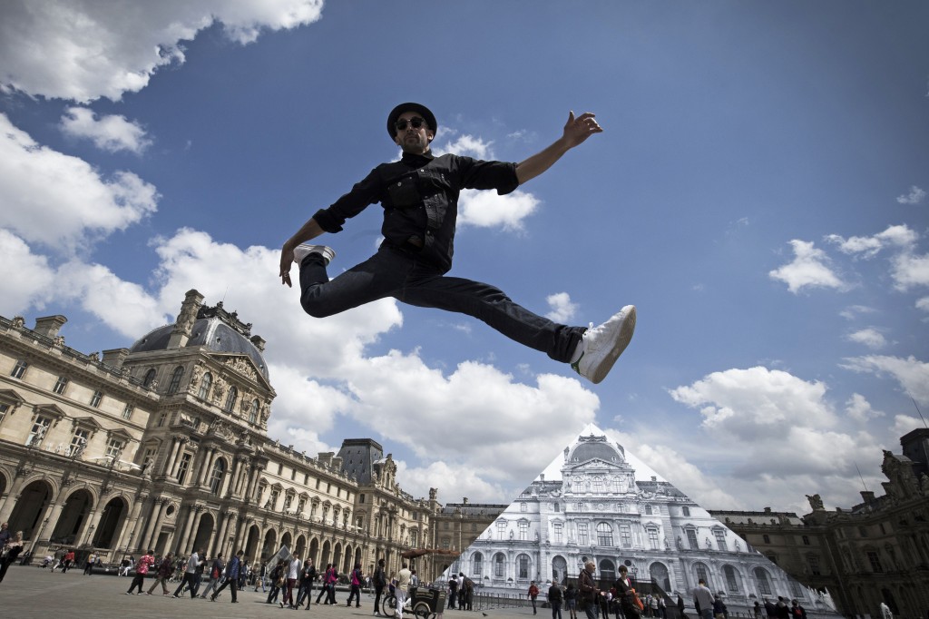 French artist and photographer JR jumps in front the Louvre pyramid after recovering it 'with a surprising anamorphic image', according to the museum in Paris on May 25, 2016.  This recovering introduces the opening of JR exhibition 'Contemporary art  JR at the Louvre' will run from May 25 to June 27, 2016.  / AFP PHOTO / JOEL SAGET