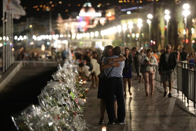 Two people hold each other by the new makeshift memorial on July 18, 2016 in Nice, in tribute to the victims of the deadly Bastille Day attack at the Promenade des Anglais after it was moved from the pavement of the road to the seafront so that the street can be re-opened. France was set to hold a minute's silence on July 18, 2016 to honour the 84 victims of Mohamed Lahouaiej-Bouhlel, a 31-year-old Tunisian who drove a truck into a crowd watching a fireworks display on Bastille Day, but a period of national mourning was overshadowed by bickering politicians. Church bells will toll across the country, and the country will fall silent at midday, a now grimly familiar ritual after the third major terror attack in 18 months on French soil. / AFP PHOTO / Valery HACHE
