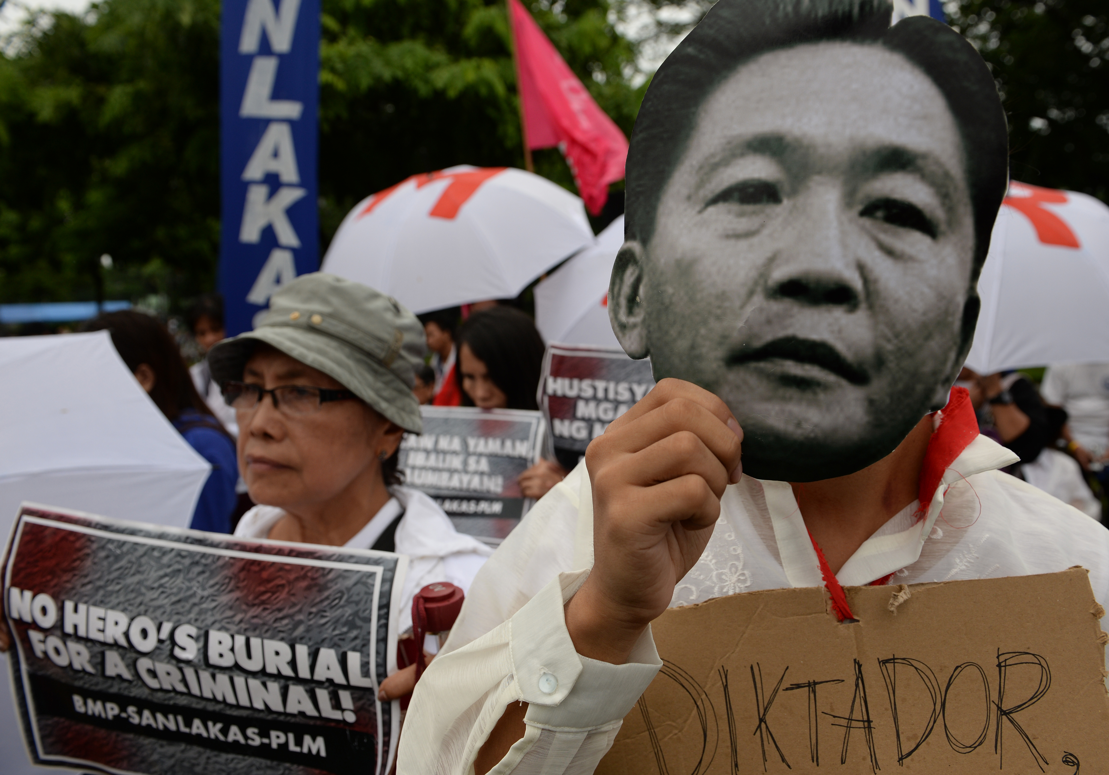 A protester (R) holds a mask featuring a portrait of the late dictator Ferdinand Marcos, while another (L) displays a placard with an anti-Marcos slogan during a demonstration at a park in Manila on August 14, 2016, against plans to honour the late dictator Ferdinand Marcos with a state burial. About 2,000 people gathered in heavy rain to denounce Philippine President Rodrigo Duterte's plans to move late dictator Ferdinand Marcos' remains from his northern hometown to the National Heroes' Cemetery in the capital, Manila, next month. / AFP PHOTO / TED ALJIBE