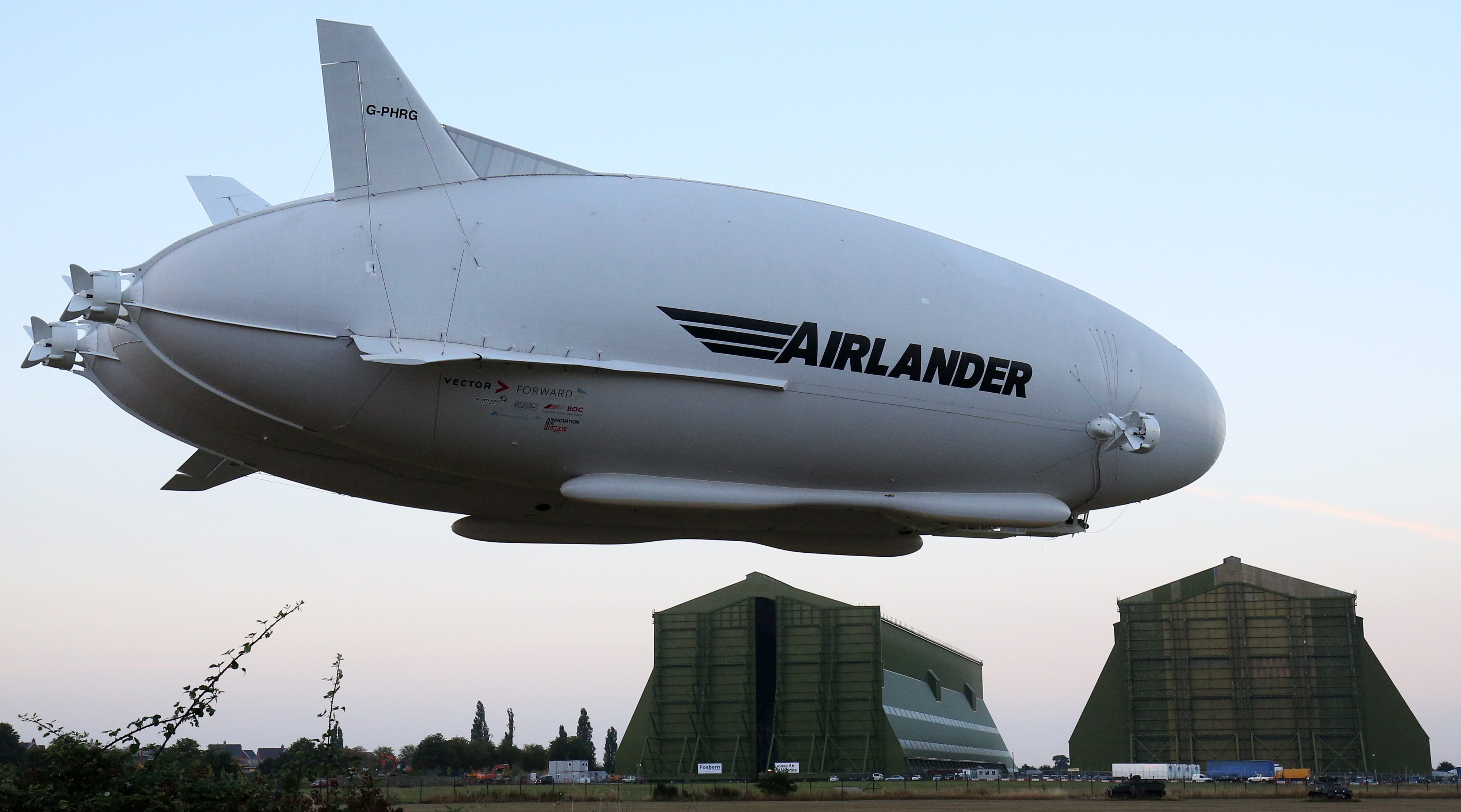 The Hybrid Air Vehicles HAV 304 Airlander 10 hybrid airship is seen with hangars in the background on its maiden flight at Cardington Airfield near Bedford, north of London, on August 17, 2016.  The Hybrid Air Vehicles 92-metre long, 43.5-metre wide Airlander 10, billed as the world's longest aircraft, lifted off for the first time from an airfield north of London. The Airlander 10 has a large helium-filled fabric hull and is propelled by four turbocharged diesel engines. According to the company it can stay airborne for up to five days at a time if manned, and for over 2 weeks unmanned with a cruising speed of just under 150 km per hour and a payload capacity of up to 10,000 kg. / AFP PHOTO / JUSTIN TALLIS
