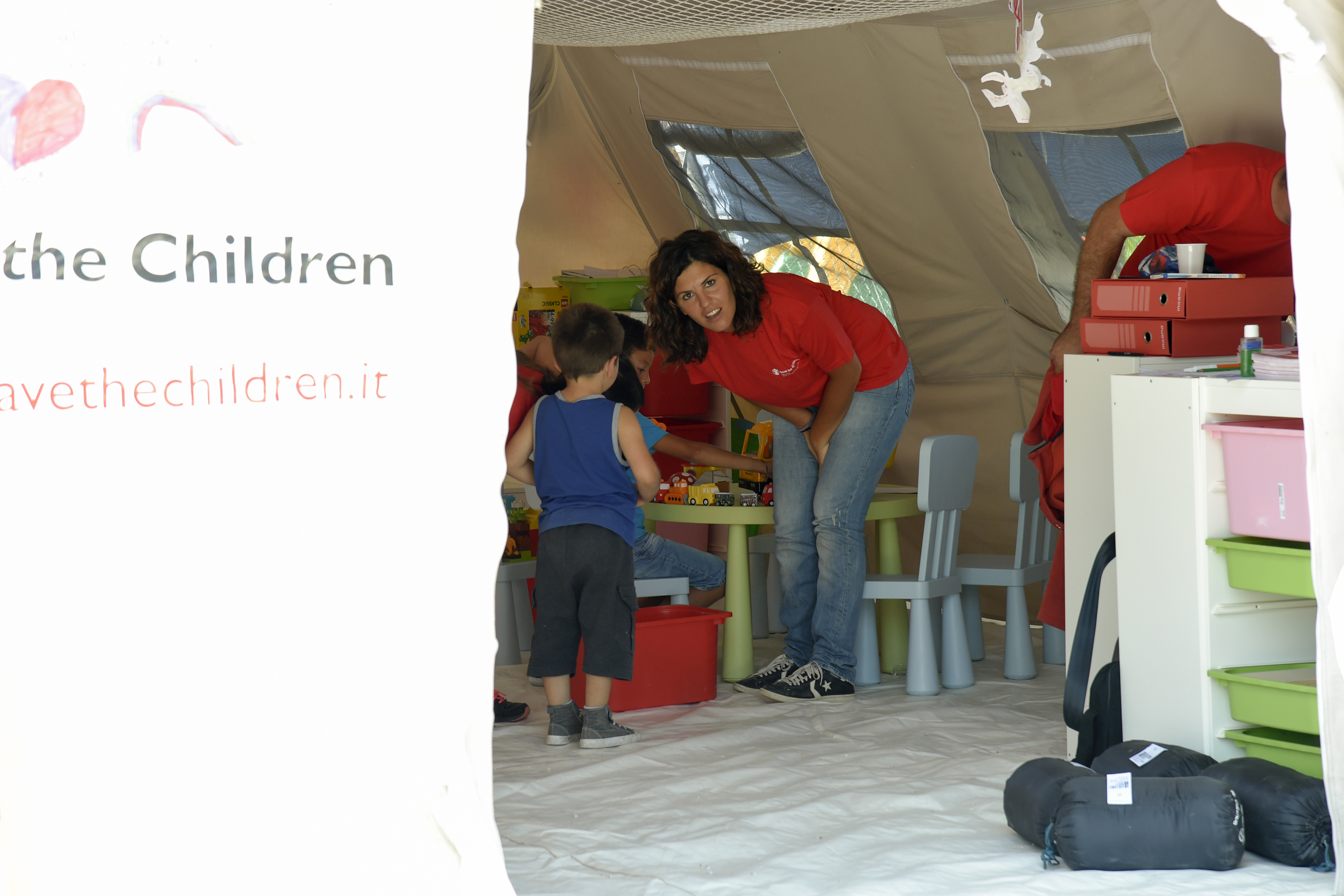 A member of NGO Save the Children stands next to a child in a tent inside a camp at the damaged central Italian village of Amatrice on August 27, 2016, three days after a 6.2-magnitude earthquake struck the region killing  some 281 people. Italy prepared for an emotional day of mourning on August 27, 2016 with flags across the country to fly at half mast in honour of the 281 victims of a devastating earthquake. Grieving families began burying their dead on August 26 as rescue workers combing the rubble said they had found no new survivors in the remote mountain villages in central Italy blitzed by August 24's powerful pre-dawn quake.  / AFP PHOTO / ANDREAS SOLARO