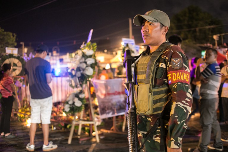 A military personnel stands guard as residents place flowers and lighted candles at a blast site in Davao City, in the southern island of Mindanao on September 4, 2016, as authorities tightened its security in public places two days after a bomb explosion at a night market that left 14 people dead and 60 wounded. Philippines police September 4 were searching for three people wanted for questioning over the bombing of a night market in President Rodrigo Duterte's hometown blamed on an Islamic militant group. / AFP PHOTO / MANMAN DEJETO