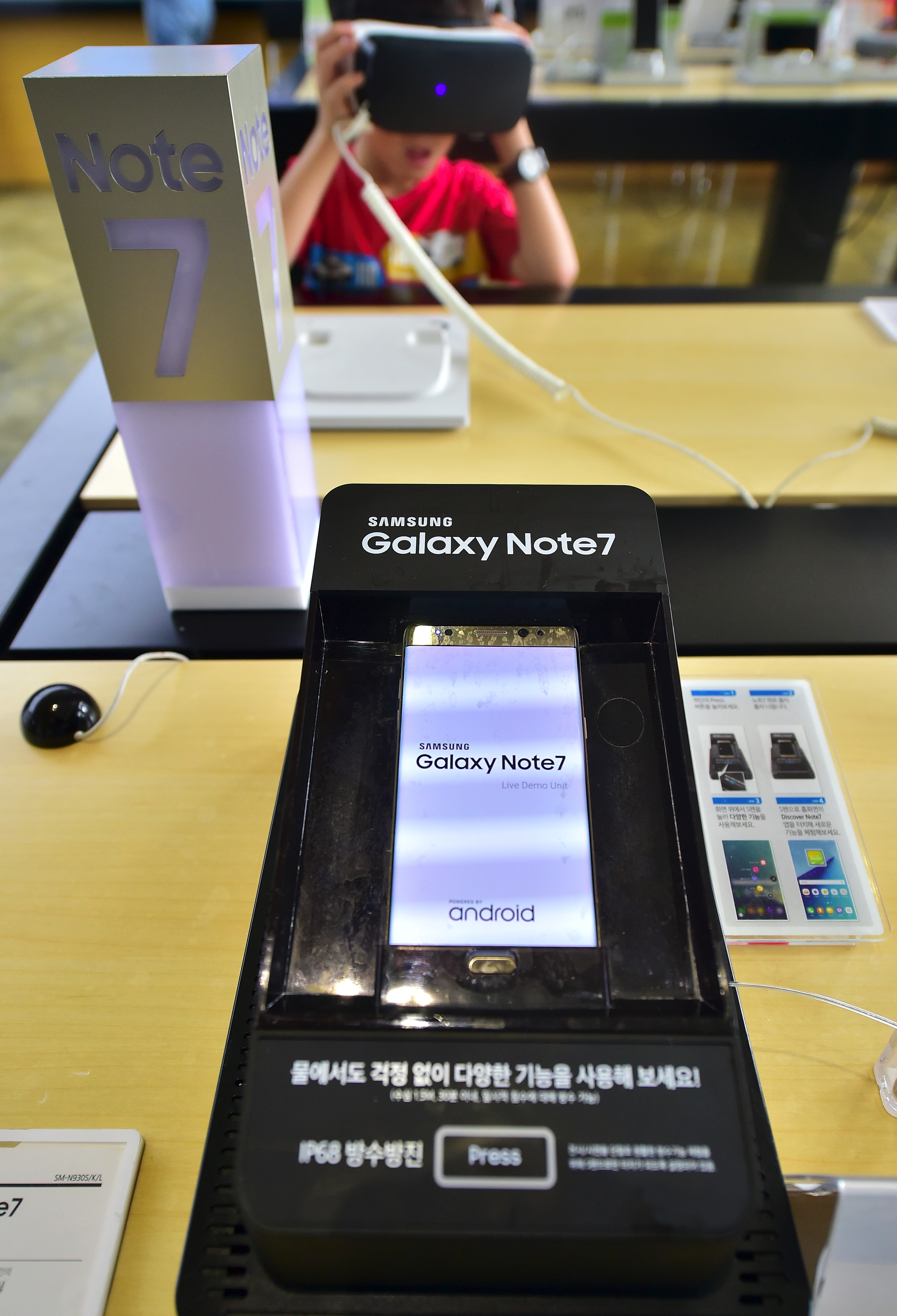 A Galaxy Note 7 smartphone is on display in a waterproof-testing case, whilst a boy (back) experiences Samsung Gear VR at a mobile phone shop in Seoul on September 12, 2016. Samsung shares plunged on September 12 after the South Korean electronics giant urged global users to stop using its Galaxy Note 7 smartphone due to a spate of exploding batteries that raised alarm around the world. / AFP PHOTO / JUNG YEON-JE