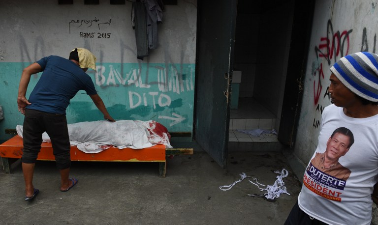 A man wearing an election campaign of then presidential candidate and now President Rodrigo Duterte, stands next to cadaver of a Muslim man wrapped in clothes, killed during a illegal drug buy-bust police operation in Quiapo district of Manila City on September 16, 2016. The Philippines faced calls on September 16 to investigate its firebrand president after a self-confessed hitman tagged the Philippine president in alleged extrajudicial killings in Davao City when he was still a city mayor. / AFP PHOTO / TED ALJIBE