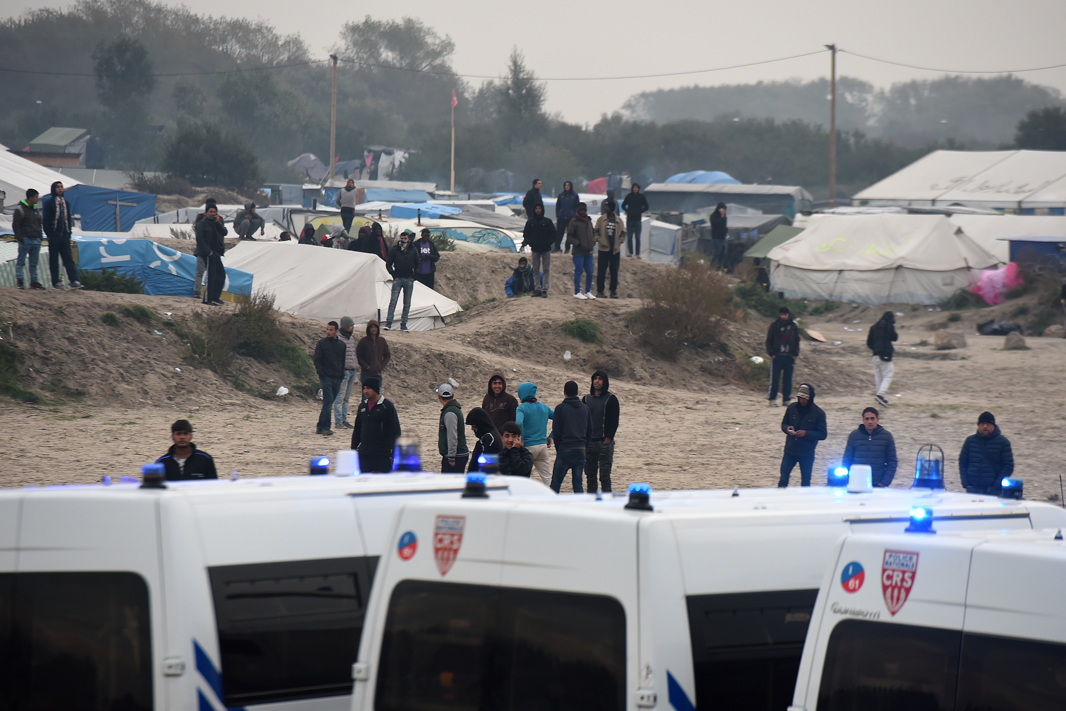 Migrants gather near police vehicles at the "Jungle" migrant camp in Calais, northern France, on October 23, 2016 on the eve of the camp's planned evacuation. On the eve of the demolition of the Calais "Jungle" camp, French officials handed out flyers in several languages notifying migrants of the camp's imminent closure and urging them to abandon their dreams of reaching Britain. / AFP PHOTO / DENIS CHARLET